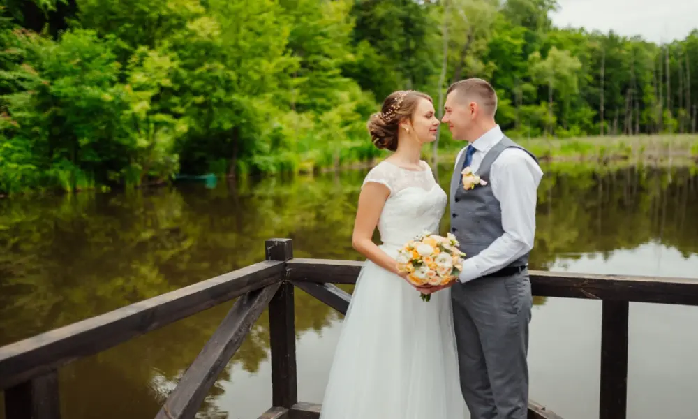 A bride and groom stand close together on a wooden deck by a calm lake at one of the beautiful wedding venues in Greater Manchester, surrounded by lush green trees. The bride holds a bouquet and wears a white dress; the groom is in a gray vest and trousers.