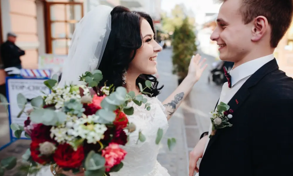 A bride in a white dress and veil smiles joyfully at the groom, holding a bouquet of red and white flowers, while the groom, dressed in a dark suit, smiles back outdoors near popular wedding venues in Greater Manchester.