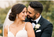 A bride in a white lace dress and veil smiles lovingly as a groom in a black tuxedo with a white rose boutonniere embraces her outdoors.
