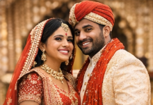 A bride and groom in traditional Indian wedding attire smile together. The bride wears a red lehenga with gold jewelry, and the groom wears a cream sherwani with a red turban and scarf. Ornate decorations are in the background.