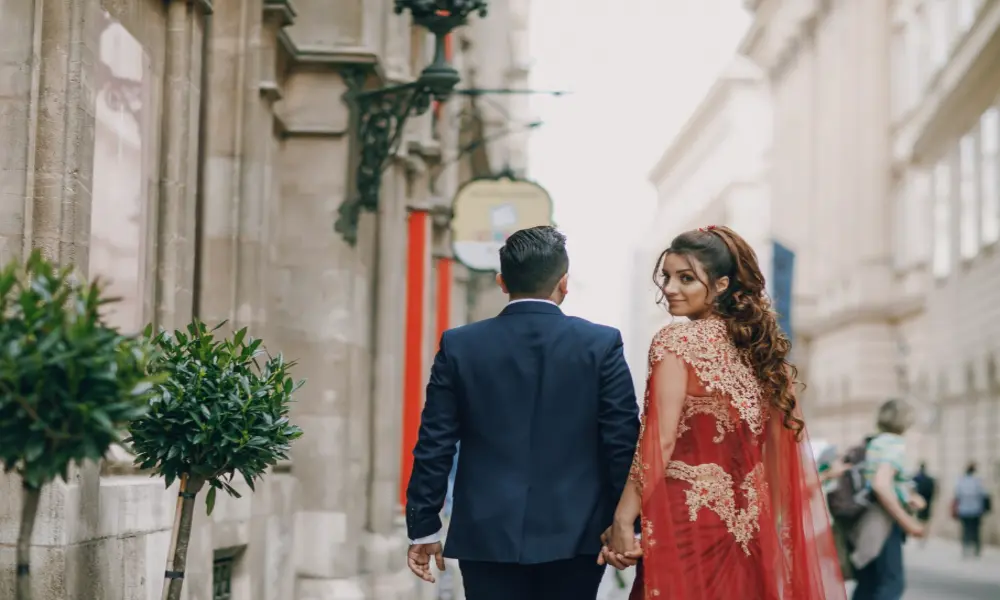 A couple walks hand in hand down a city street; the woman wears an ornate red and gold dress and looks back over her shoulder while the man, in a dark suit, faces forward.