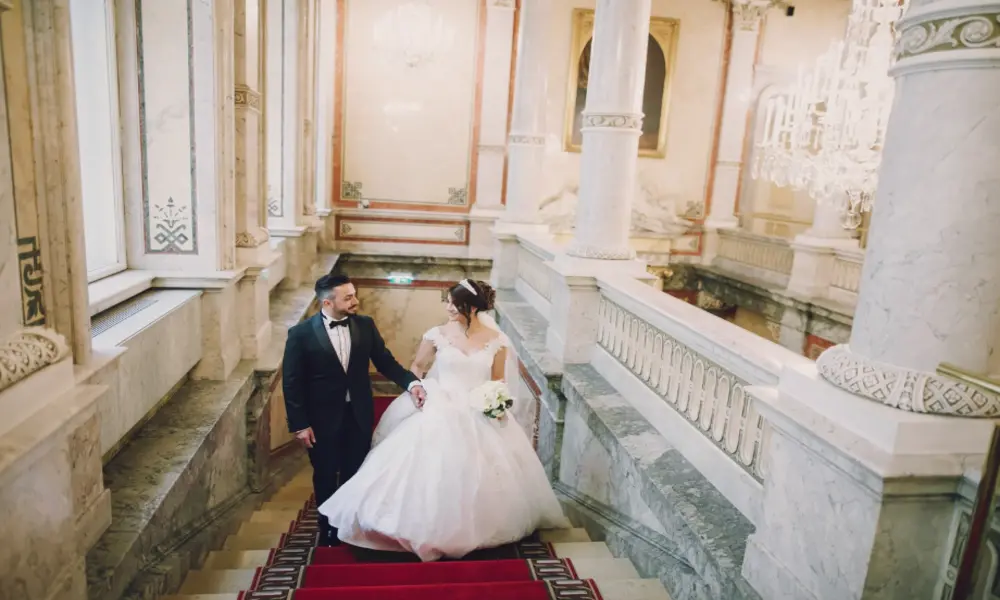 A bride in a white gown and a groom in a black tuxedo hold hands while walking up an elegant marble staircase with red carpet in a grand, ornate hall—perfect inspiration for your Greater Manchester wedding.