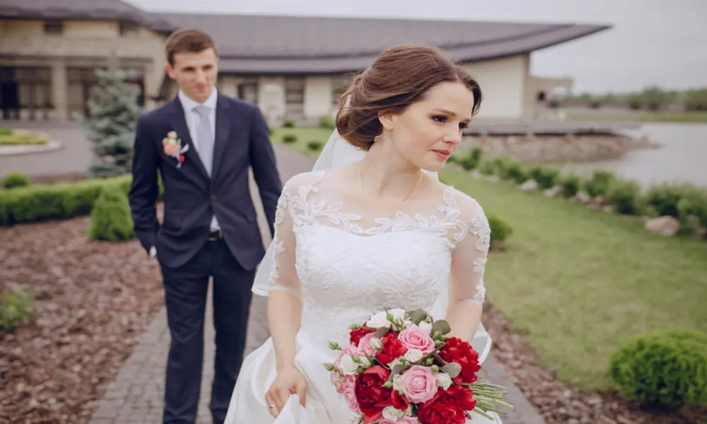 A bride in a white dress holding a bouquet of red and pink flowers stands outdoors, while a groom in a dark suit stands behind her on a garden path near a building—a picturesque scene perfect for any 2026 wedding guide or Greater Manchester wedding.
