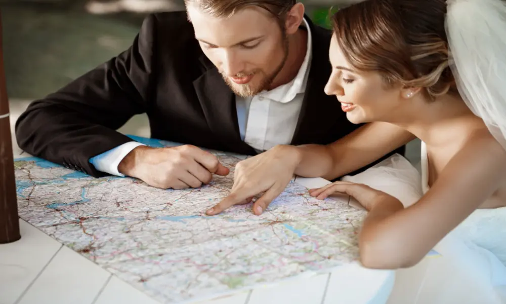 A bride and groom in wedding attire sit together at a table, looking closely at and pointing to locations on a large paper map as they plan their search for the perfect Greater Manchester wedding venues.