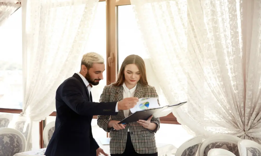 A man and a woman in business attire stand by a window with lace curtains, reviewing a folder of colorful charts—perhaps planning Greater Manchester wedding venues for a 2026 wedding guide. The man points at the document while the woman holds it and listens.